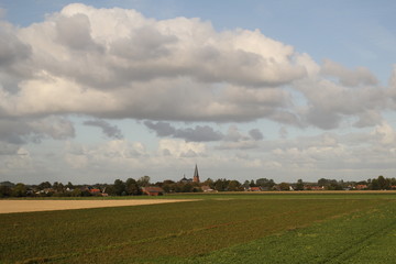 a landscape in the dutch countryside in springtime with a little village with a church behind the fields with growing green crops and a blue sky with big white clouds