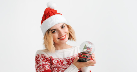Young blond woman in red cap Christmas costume smiles and looks at Christmas Snow Globe Santa Claus on a white background. Christmas symbol. Noel. Copy space. Christmas preparation. Emotions