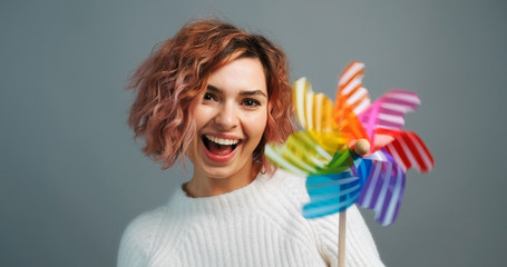 Portrait happy young woman holds windmill and rotates hand smilingly looking at camera, rotating moment of the mill on grey background. Colors of LGBT flag. Freedom of choice. Lifestyle