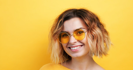 Portrait of happy young woman correcting yellow glasses on her forehead coquettishly showing tongue to camera smiling on an isolated yellow background. Monotone. Emotions of people. Copy space