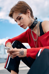 Urban Style. Young stylish woman sitting outdoors browsing internet on smartphone concentrated...
