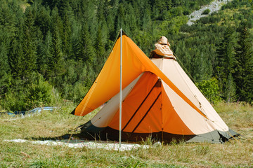 Tourist orange tent among mountains and a pine forest in summer on meadow with green grass at campsite sunny day. Family holidays with tent. Relax. Travel © marina_larina
