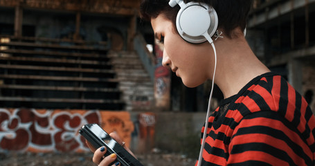 Asian teenager girl listens to favorite music on headphones from the phone, holding it in her hands against the background of an abandoned old unfinished building. Hipster, freak, emo, grunge