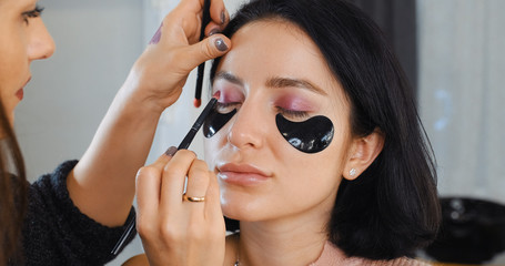 Portrait of brunette girl with patches on her face in makeup studio, makeup artist applies eye shadow with brush dipping into palette against background of bokeh light bulbs mirror. Makeup and beauty