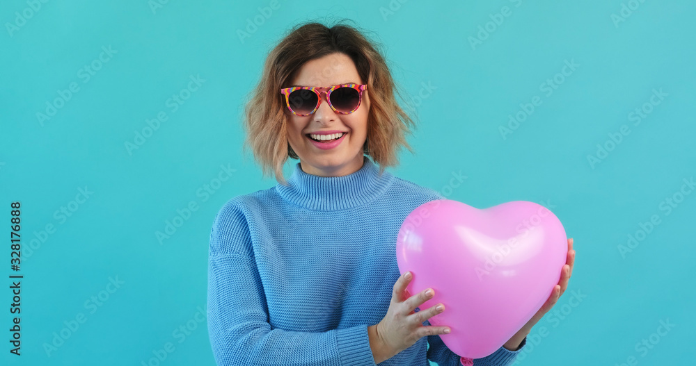 Wall mural Valentine's Day cheerful girl pink heart balloon rhythmic moves from himself, smiling happily bright blue background looking at camera. Woman, symbol of love. Holiday of all lovers. Feelings, emotion