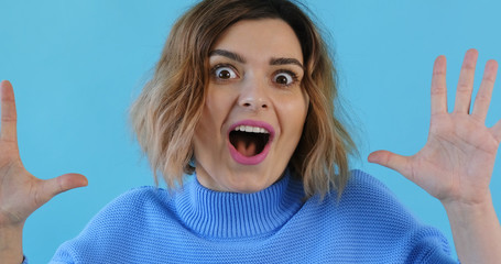 Portrait of happy young sexy girl in blue sweater, stands holding her hands up in surprise, covering her mouth on blue background. Pleasure. The emotions of people. Monotone. Copy space. Blue pantone