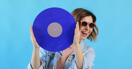 Portrait of happy young sexy girl who hides behind blue vinyl record for turntable and appears on right  in frame, smiles looks at camera on blue background. Emotions people. Monotone. Blue pantone