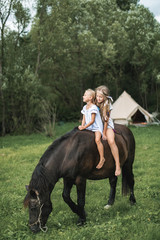 Two pretty little girls in casual boho clothes galloping on beautiful dark horse outdoors in the field. Green trees and white teepee wigwam tent on the background