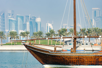 Retro historical boat with blurred panoramic view of modern skyline of Doha and green palms on background