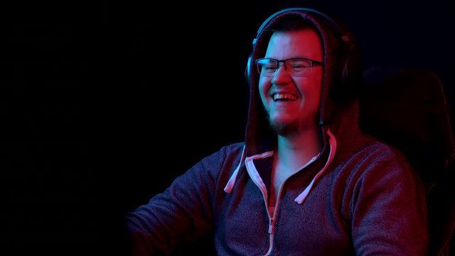 A young man sits at a computer with headphones. A man in a hood looks at a computer monitor and laughs. The camera zooms out from behind the monitor.