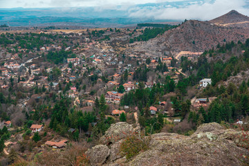 View from a mountain, the city of La Cumbrecita surrounded by small elevations ands trees, mainly evergreens. In the back a mountain range with a few clouds on the sky