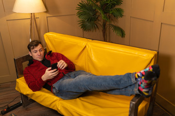 Obraz premium A confident, happy young man is resting on the couch at home. A visiting man in casual clothes while sitting with a smartphone on a yellow sofa at home