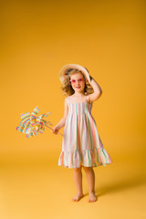 child girl blonde smiling in sunglasses and summer sundress holding a windmill stands on a yellow background. happy child. space for text