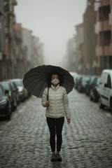 Pandemic. A mixed-race woman in antiviral mask stands in the street in cloudy weather.