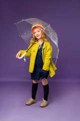 child girl blonde smiling in a green coat and colorful hat holding an umbrella. purple background, space for text