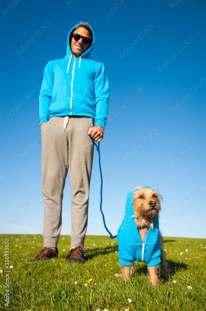 Wall mural Small dog standing outdoors with his best friend owner in matching blue hoodies on bright green field