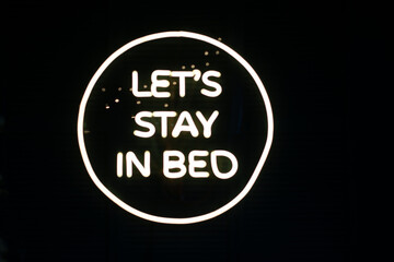 Fototapeta na wymiar Phrase Let's stay in bed written in shiny neon letters on black background