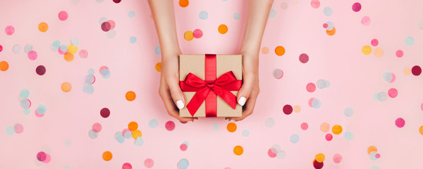 Banner. Woman hands holding present box with red bow on pastel pink background with multicolored confetti. Flat lay style. - 328186645