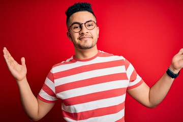 Young handsome man wearing casual striped t-shirt and glasses over isolated red background clueless and confused expression with arms and hands raised. Doubt concept.
