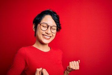 Naklejka premium Young beautiful asian girl wearing casual t-shirt and glasses over isolated red background excited for success with arms raised and eyes closed celebrating victory smiling. Winner concept.