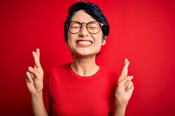 Young beautiful asian girl wearing casual t-shirt and glasses over isolated red background gesturing finger crossed smiling with hope and eyes closed. Luck and superstitious concept.