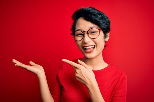 Young beautiful asian girl wearing casual t-shirt and glasses over isolated red background amazed and smiling to the camera while presenting with hand and pointing with finger.