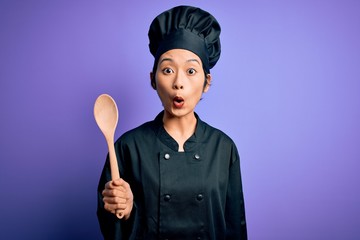 Young beautiful chinese chef woman wearing cooker uniform and hat holding wooden spoon scared in...