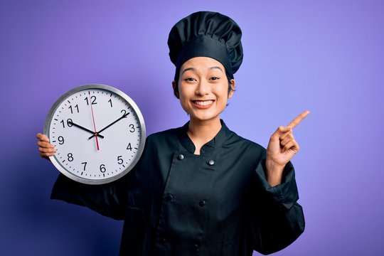 Young chinese chef woman wearing cooker uniform and hat holding clock doing countdown surprised with an idea or question pointing finger with happy face, number one