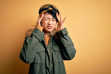 Beautiful asian skier girl wearing snow sportswear using ski goggles over yellow background with hand on head for pain in head because stress. Suffering migraine.