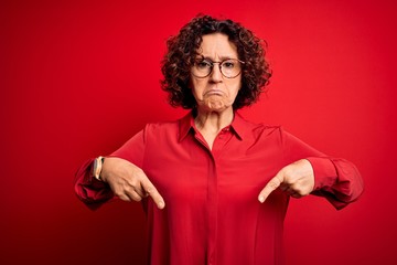 Fototapeta na wymiar Middle age beautiful curly hair woman wearing casual shirt and glasses over red background Pointing down looking sad and upset, indicating direction with fingers, unhappy and depressed.