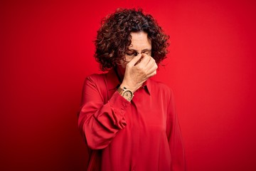 Fototapeta na wymiar Middle age beautiful curly hair woman wearing casual shirt and glasses over red background tired rubbing nose and eyes feeling fatigue and headache. Stress and frustration concept.