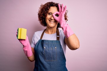 Middle age curly hair woman cleaning doing housework wearing apron and gloves using scourer with...