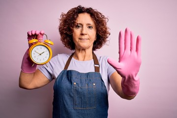 Middle age woman cleaning doing housework wearing apron and gloves holding alarm clock with open...