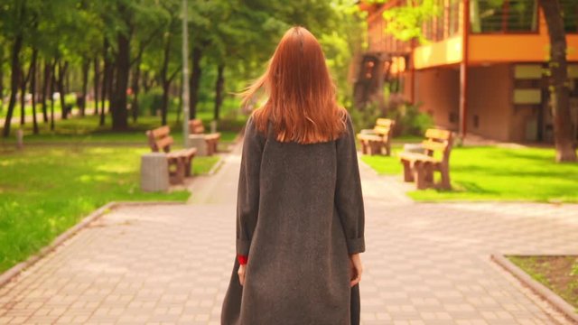 back view woman with red hair in a grey coat walks in the park with green thees enjoy sunny weather in early spring slow motion