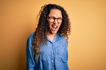 Beautiful woman with curly hair wearing casual denim shirt and glasses over yellow background...