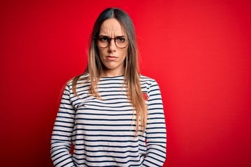 Young beautiful blonde woman with blue eyes wearing glasses standing over red background skeptic...