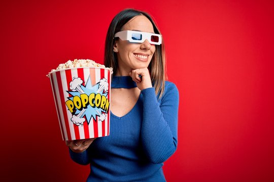 Young blonde woman wearing 3d glasses and eating pack of popcorn watching a movie on cinema with hand on chin thinking about question, pensive expression. Smiling and thoughtful face. Doubt concept.