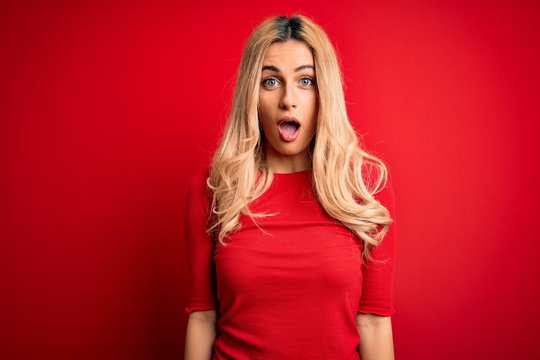 Young beautiful blonde woman wearing casual t-shirt standing over isolated red background afraid and shocked with surprise and amazed expression, fear and excited face.