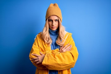 Young beautiful blonde woman wearing raincoat for rainy weather over blue background skeptic and...