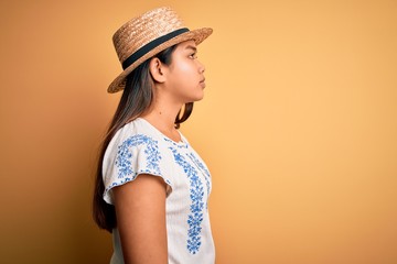 Young beautiful asian girl wearing casual t-shirt and hat standing over yellow background looking to side, relax profile pose with natural face and confident smile.