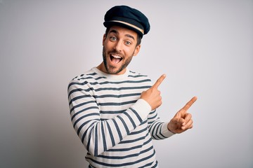 Young handsome sailor man with beard wearing navy striped uniform and captain hat smiling and looking at the camera pointing with two hands and fingers to the side. - obrazy, fototapety, plakaty