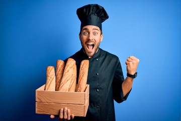 Young cooker man with beard wearing uniform holding box with bread over blue background screaming...