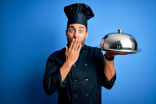 Young cooker man with beard wearing uniform holding tray with dome over blue background cover mouth with hand shocked with shame for mistake, expression of fear, scared in silence, secret concept