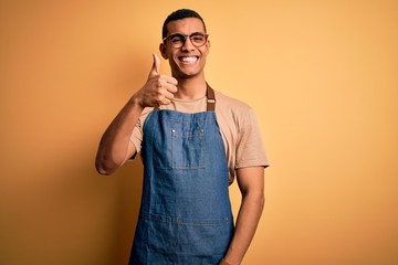 Young handsome african american shopkeeper man wearing apron over yellow background doing happy thumbs up gesture with hand. Approving expression looking at the camera showing success. - obrazy, fototapety, plakaty
