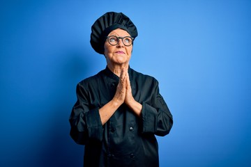 Senior beautiful grey-haired chef woman wearing cooker uniform and hat over blue background begging and praying with hands together with hope expression on face very emotional and worried. Begging.