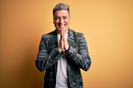 Young handsome modern man wearing business jacket standing over yellow isolated background praying with hands together asking for forgiveness smiling confident.