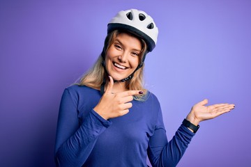 Young beautiful blonde cyclist woman wearing bike security helmet over purple background amazed and smiling to the camera while presenting with hand and pointing with finger.