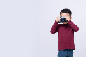 Portrait of little boy wearing red sweater standing isolated on grey background taking photo with film camera.