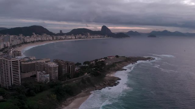 Aerial sideways pan of Copacabana fort in the foreground and Copacabana beach and Sugarloaf mountain in the background at early morning overcast sunrise in Rio de Janeiro, Brazil