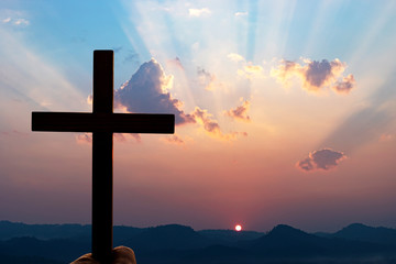 Silhouette human hands praying to the GOD while holding a crucifix symbol with bright sunbeam on the sky